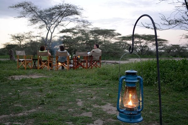 Guests in camp chairs around the campfire at dusk, a lit lantern hanging in the foreground