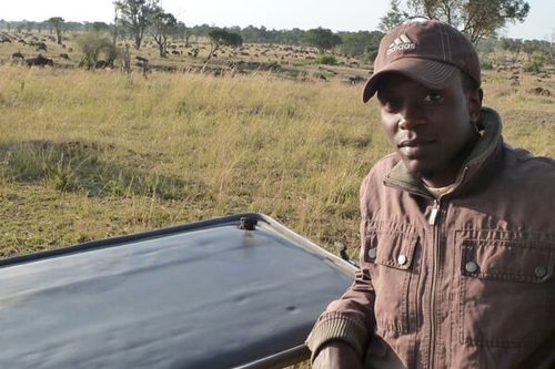 Fadhil Magoye in a cap and jacket beside a safari vehicle, a wildebeest herd on the plain behind him