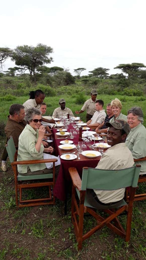 Guests and guides at a long table laid for a meal outdoors under acacia trees